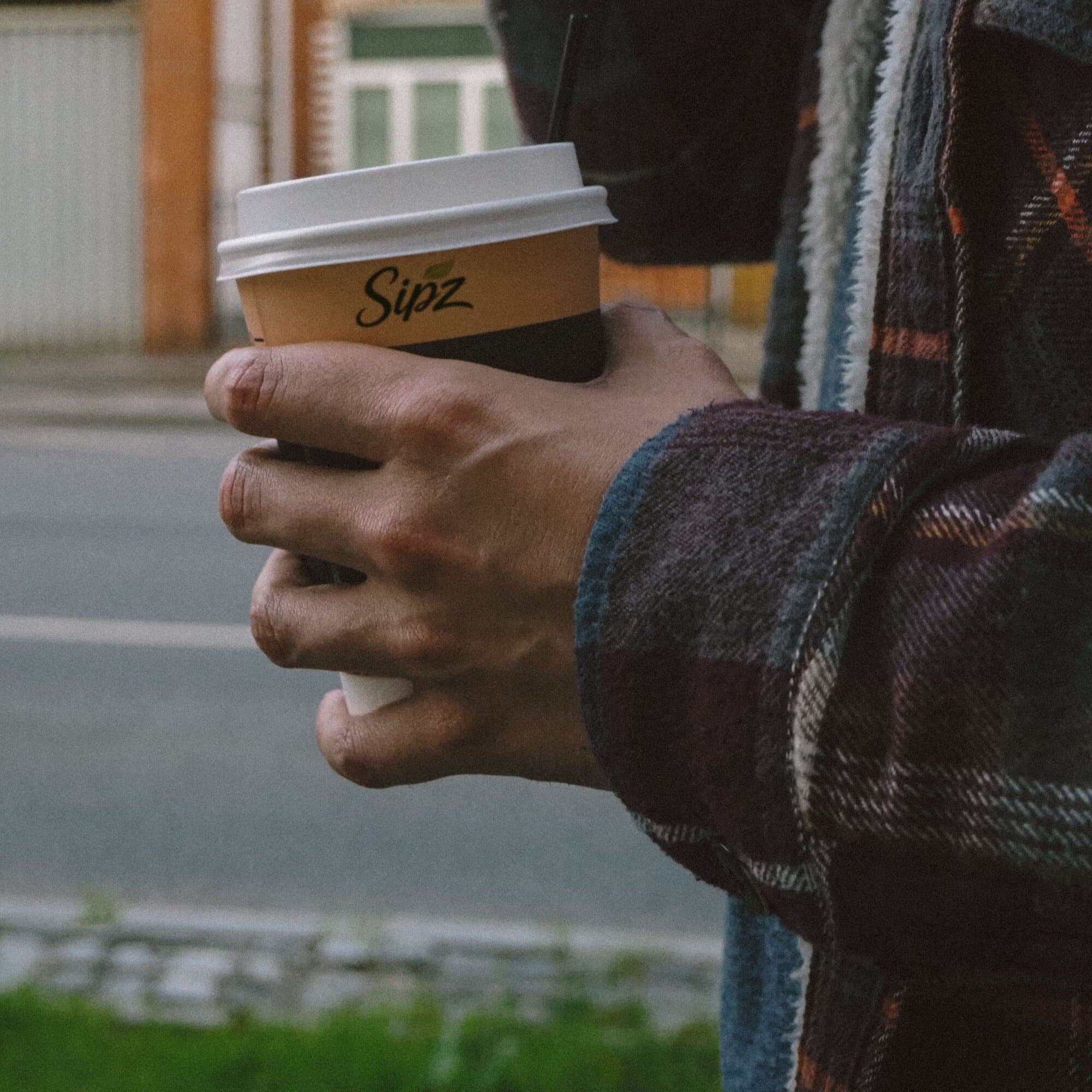A hand in a flannel sleeve holds a SIPZ coffee on a town street in the morning.