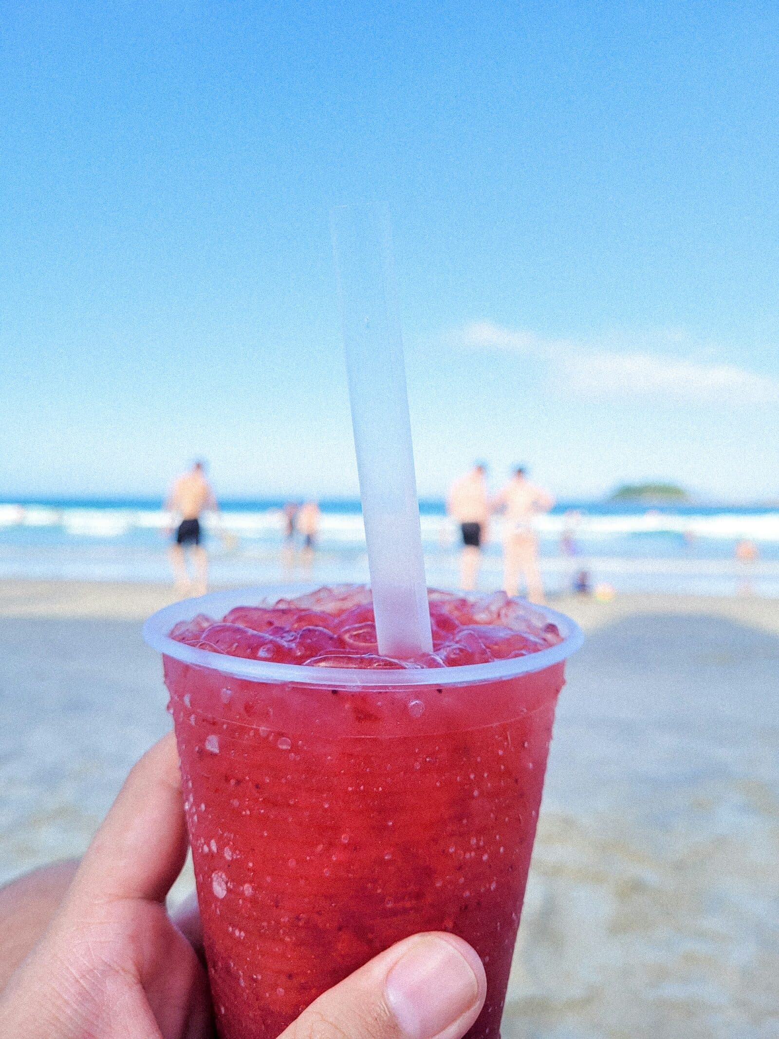 A hand holds up a red slush in a clear to-go cup on a sunny beach.